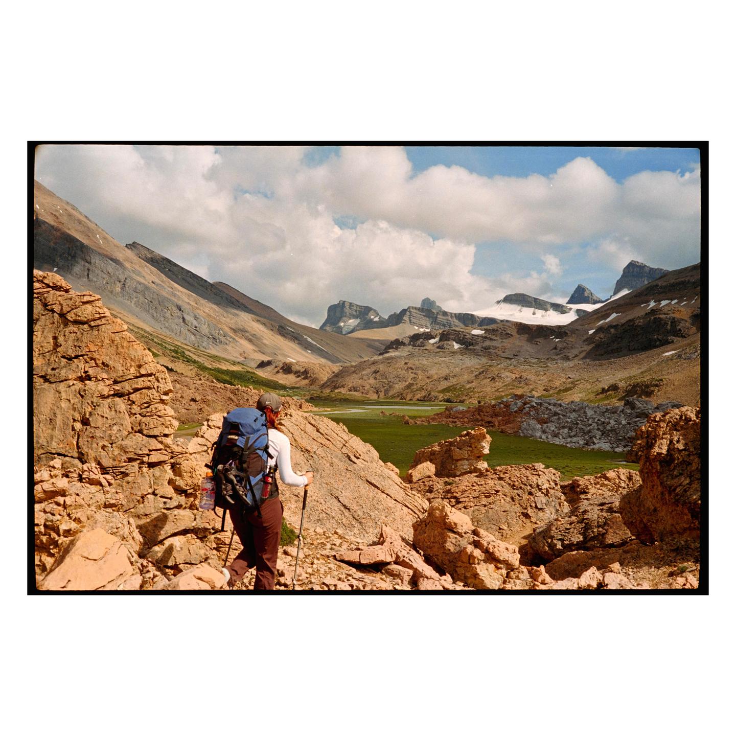 A hiker with a large backpack and trekking poles walks through a rocky mountain landscape with snow-capped peaks in the distance.