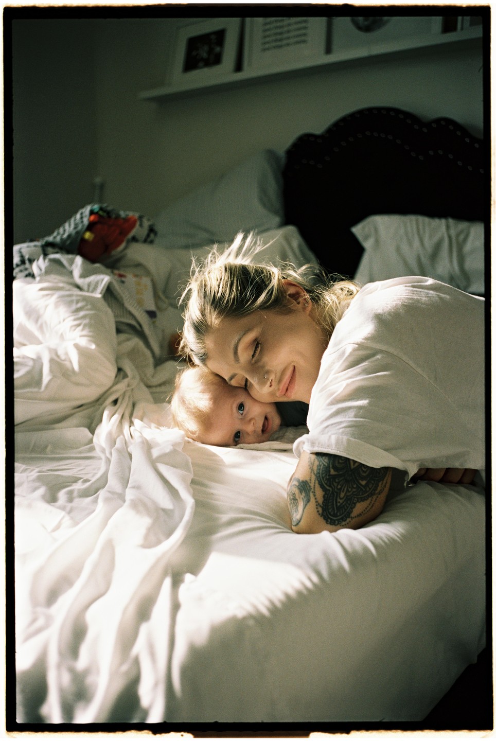 A woman with a tattoo on her arm lies on a bed next to a baby.
