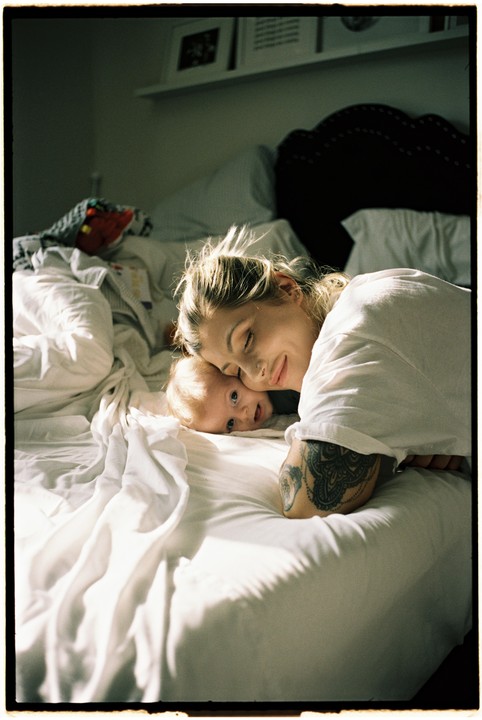 A woman with a tattoo on her arm lies on a bed next to a baby.