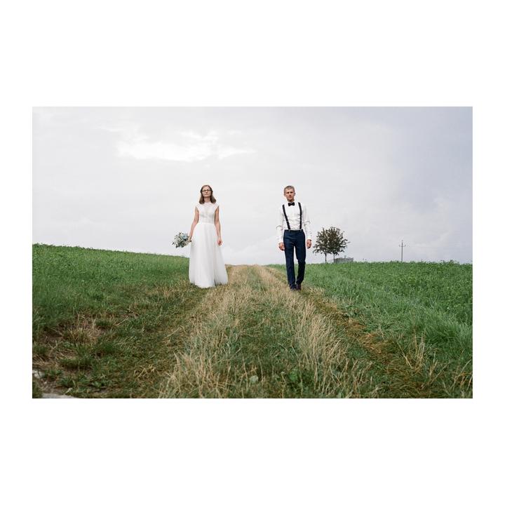 A couple walks on a dirt path through a grassy field under a cloudy sky.