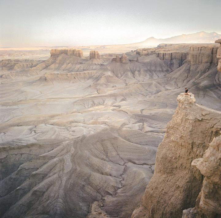 A person sits on a rock formation overlooking a vast desert landscape with mountains in the distance.