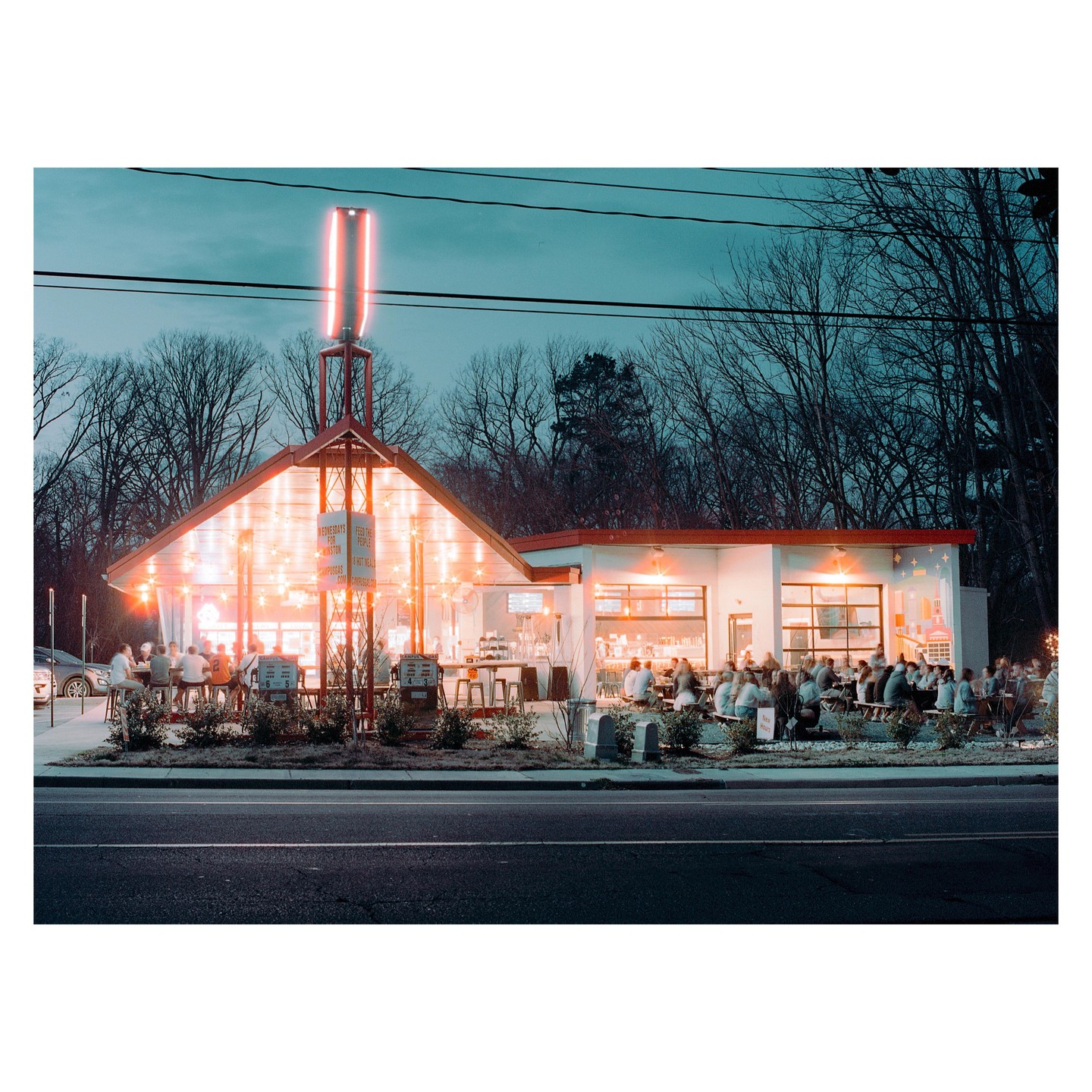 A brightly lit restaurant with many patrons sits beside a road at dusk, with bare trees in the background.