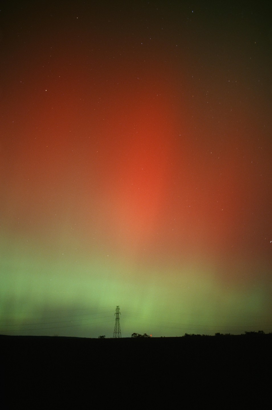 A power line tower stands silhouetted against a night sky filled with the red and green glow of the aurora borealis.