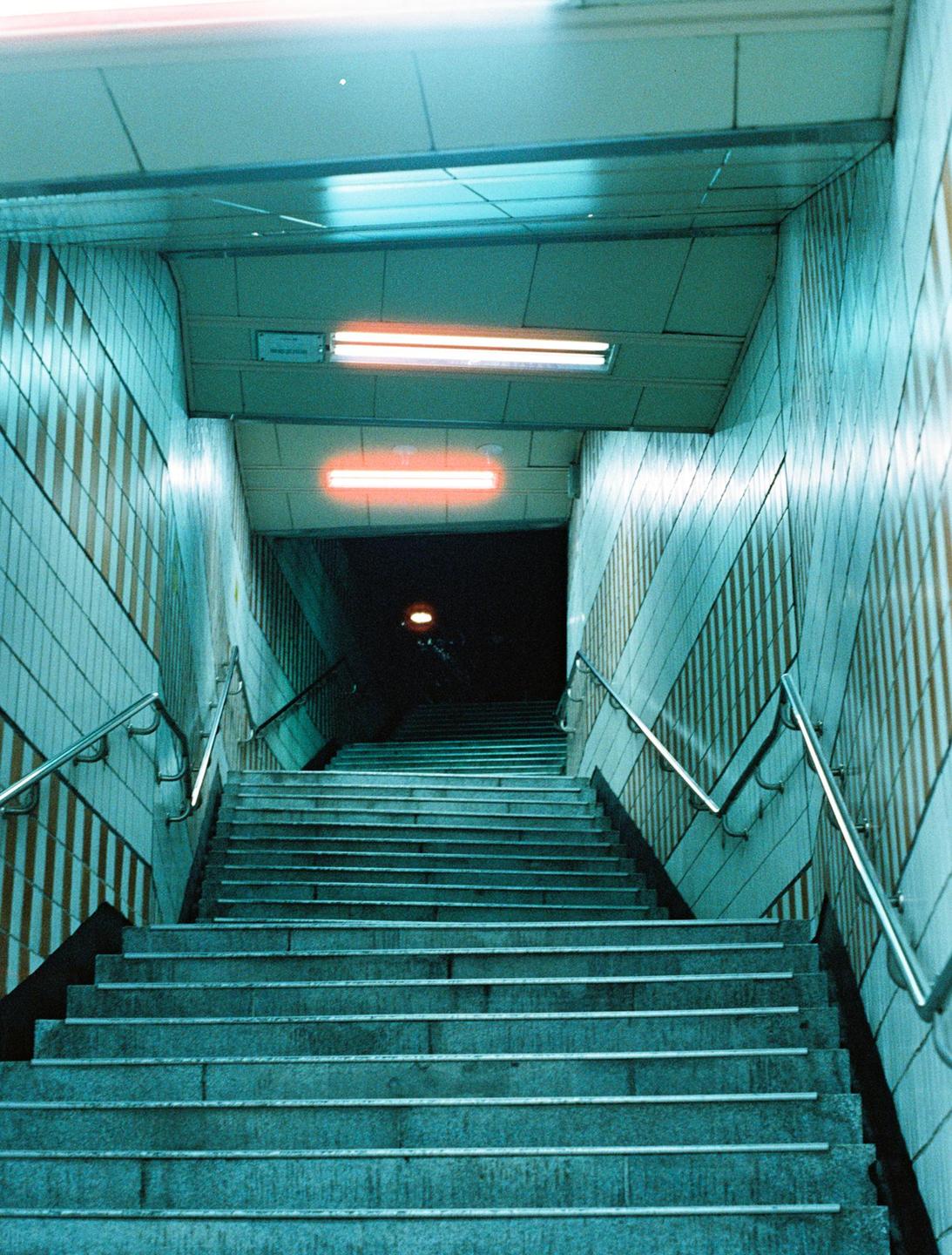 A long, tiled staircase with handrails descends into darkness, illuminated by neon lights.