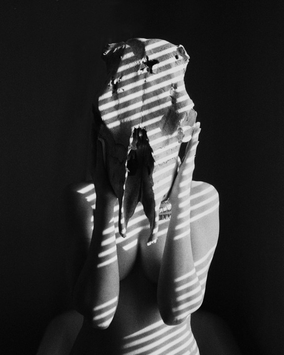 A person with a skull covering their face is illuminated by striped shadow patterns.
