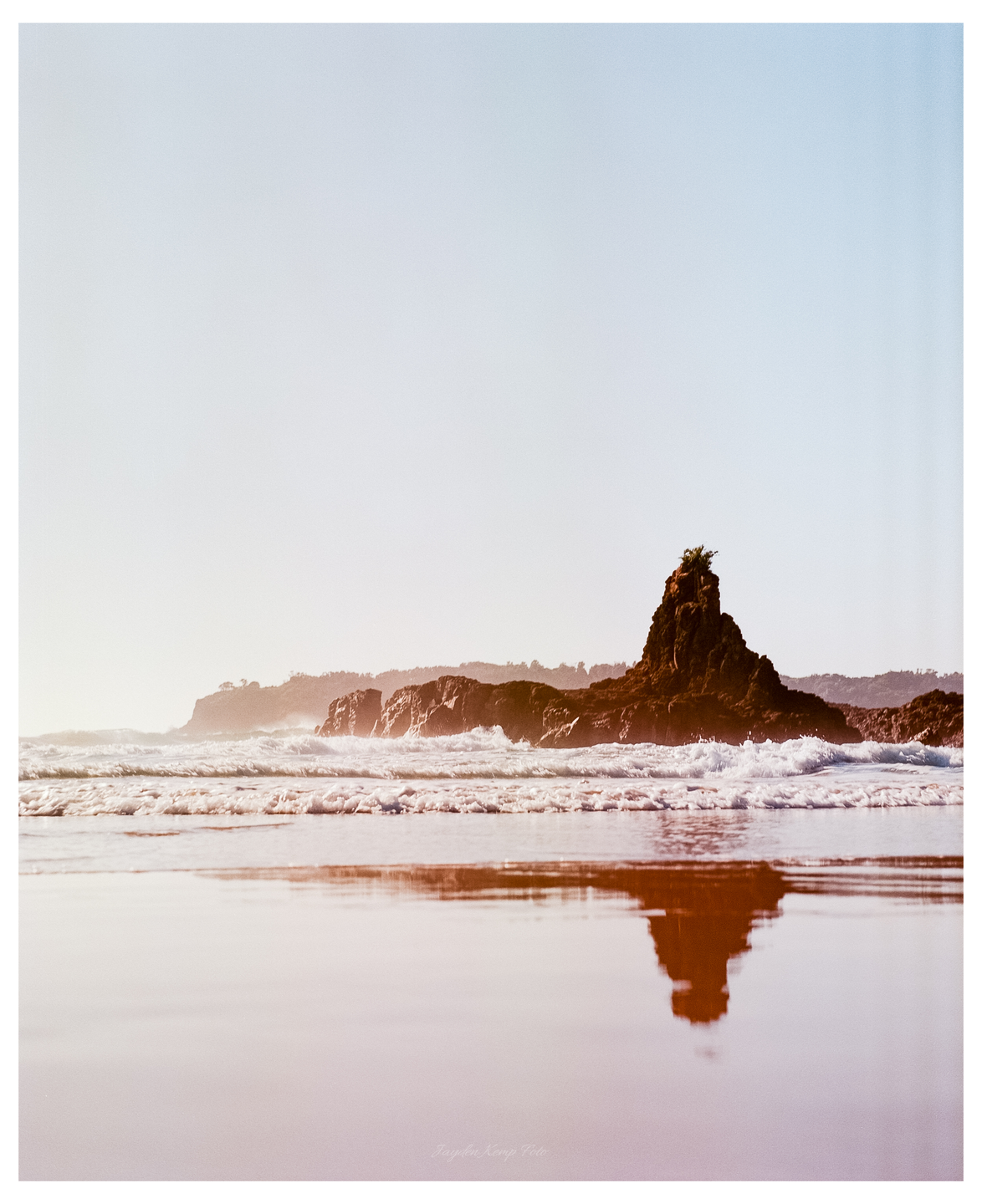 Waves crash against a rocky coast with a prominent rock formation reflecting in the wet sand.