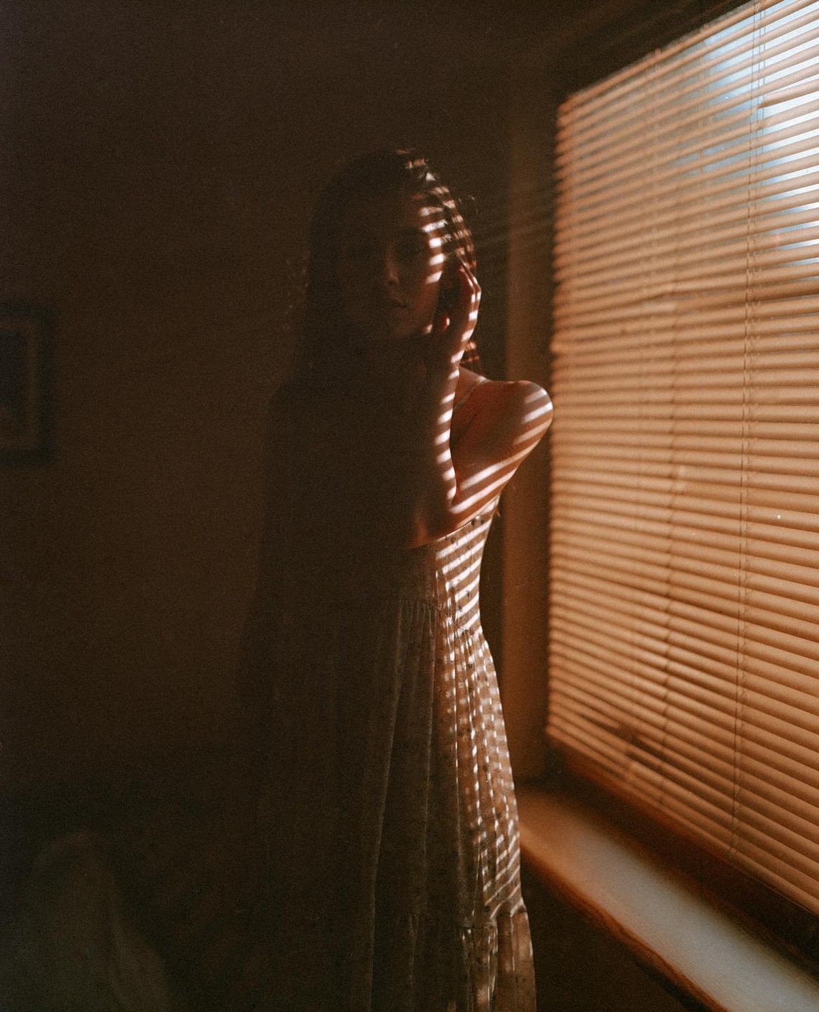 A woman stands in a dimly lit room with horizontal shadows from window blinds falling across her body.