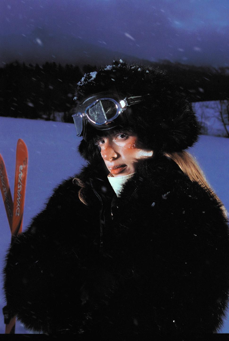 A woman wearing a fur hat and goggles stands in a snowy landscape with skis visible.