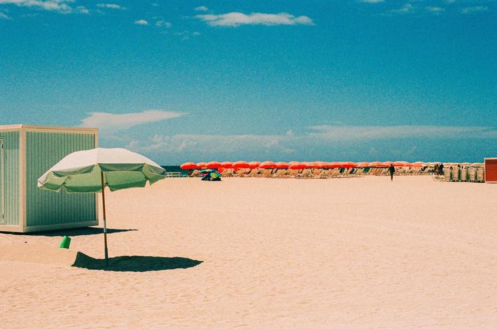 A single green beach umbrella stands in the foreground of a sandy beach with many orange umbrellas and lounge chairs in the background.