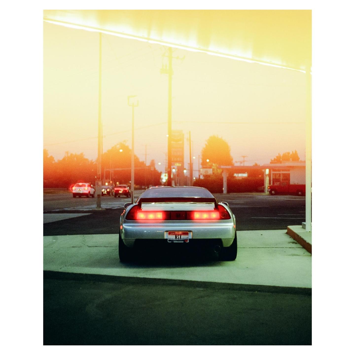 A silver sports car with illuminated taillights is parked at what appears to be a gas station during sunset.