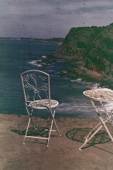 A metal chair and table sit on a cliff overlooking a rocky coastline with an ocean.