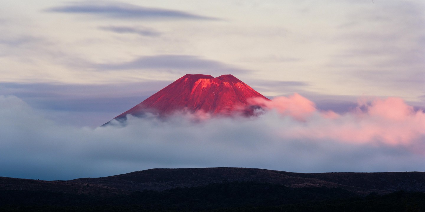 A red-hued mountain peak rises above a layer of clouds under a pastel sky.