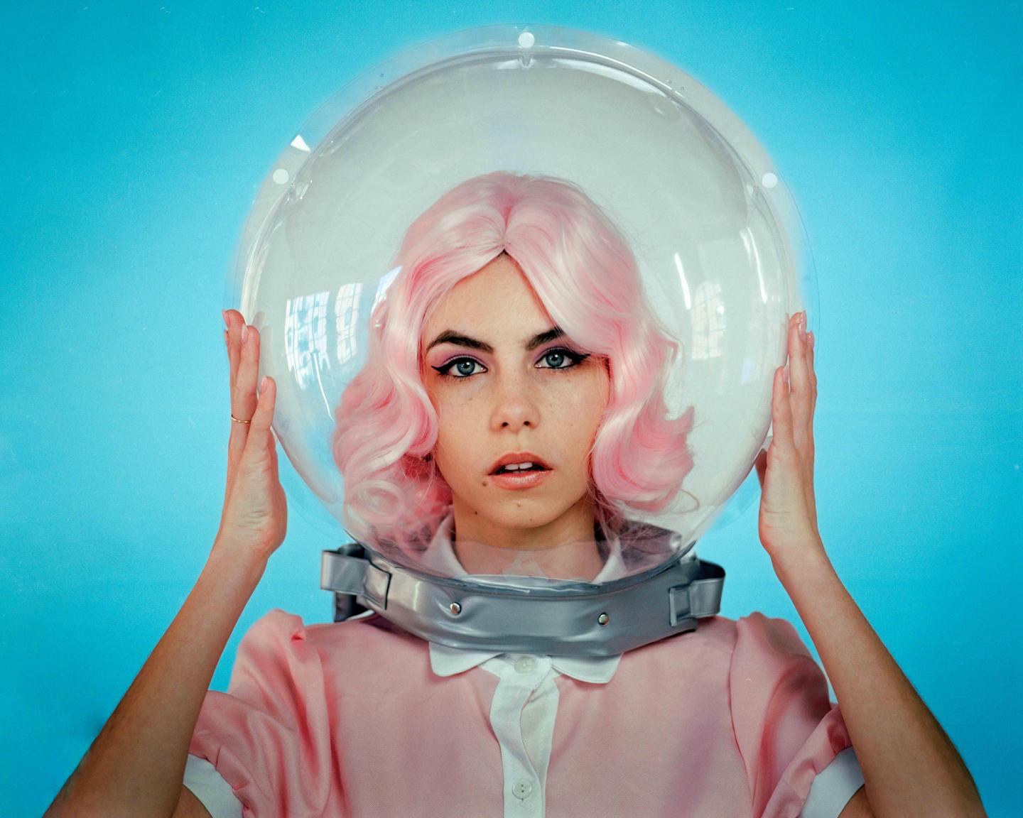 A woman with pink hair and a pink dress wears a clear space helmet against a blue background.