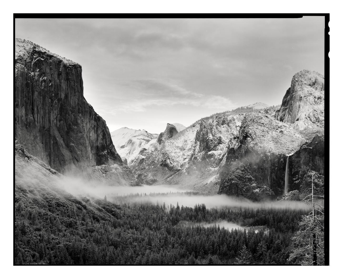 A valley with a forest and a waterfall is covered in fog and surrounded by mountains.