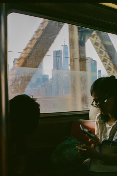 A woman looks at her phone while riding a train with a view of the New York City skyline through the window.