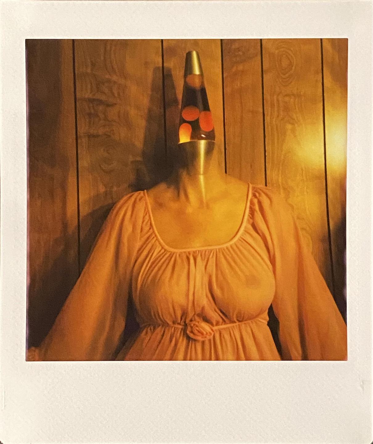 A lava lamp is placed on a woman's body, which is wearing a peach-colored dress, against a wooden wall.