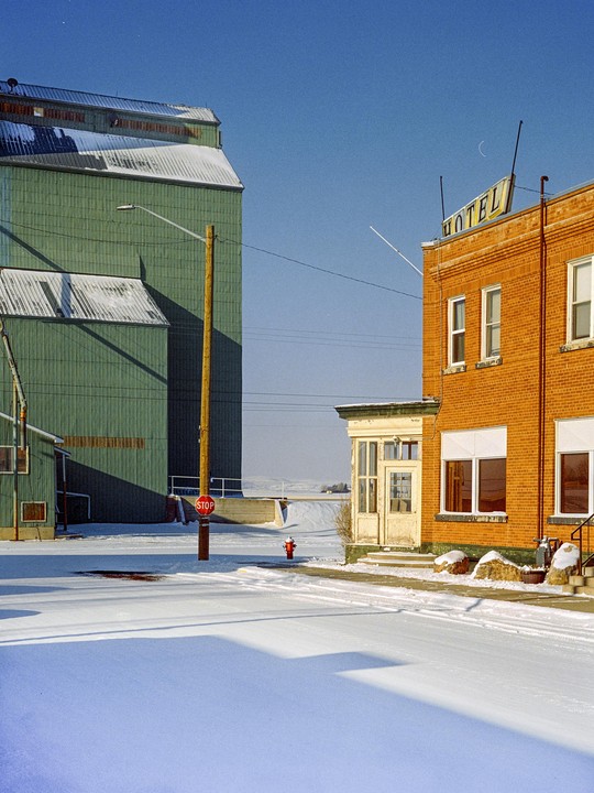 A snow-covered street is bordered by a green industrial building and a brick hotel with a "HOTEL" sign on its roof.
