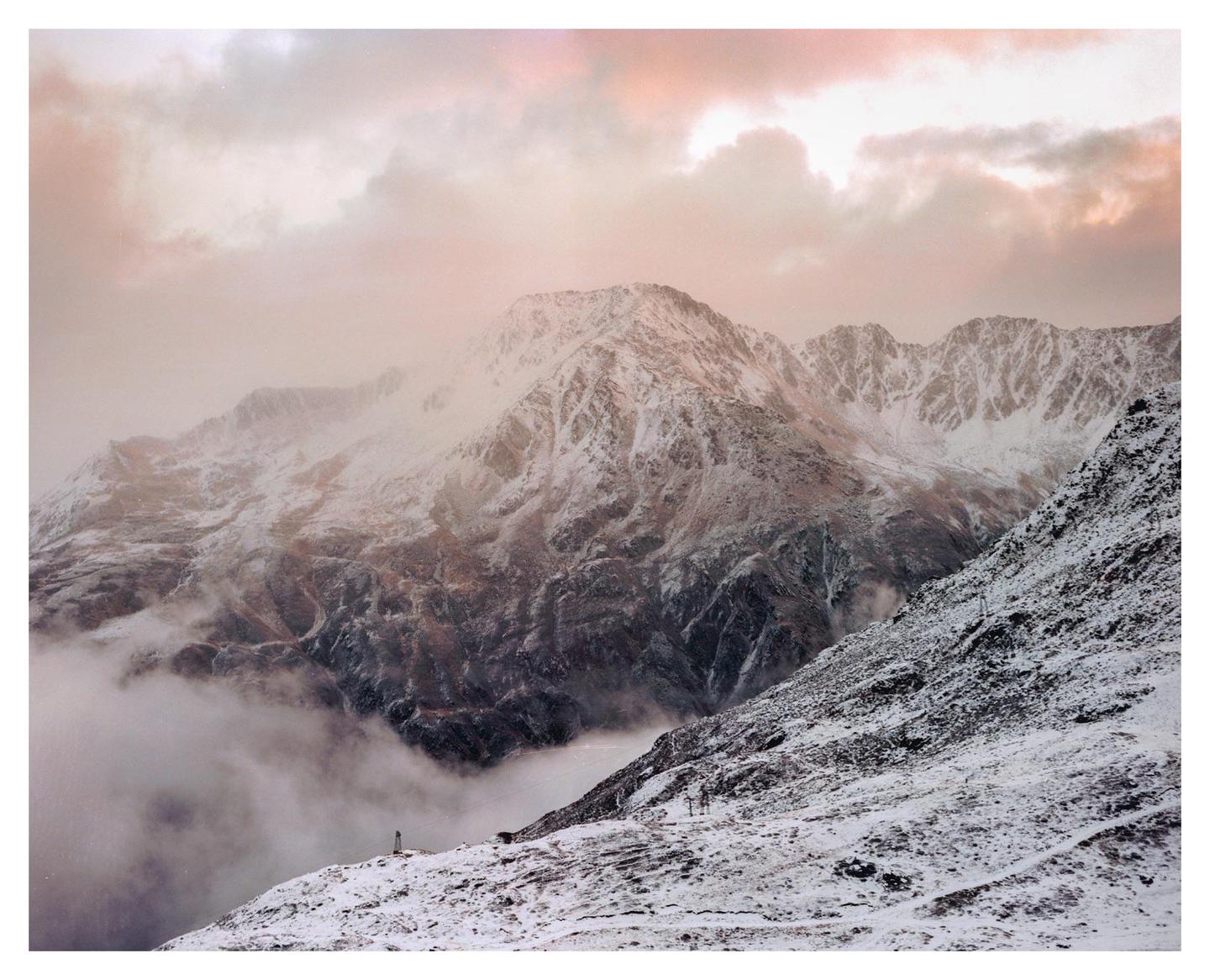 Snow-covered mountains are partially obscured by clouds under a pink sky.