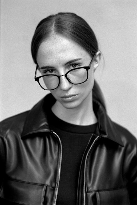 A woman wearing glasses and a leather jacket looks directly at the viewer.