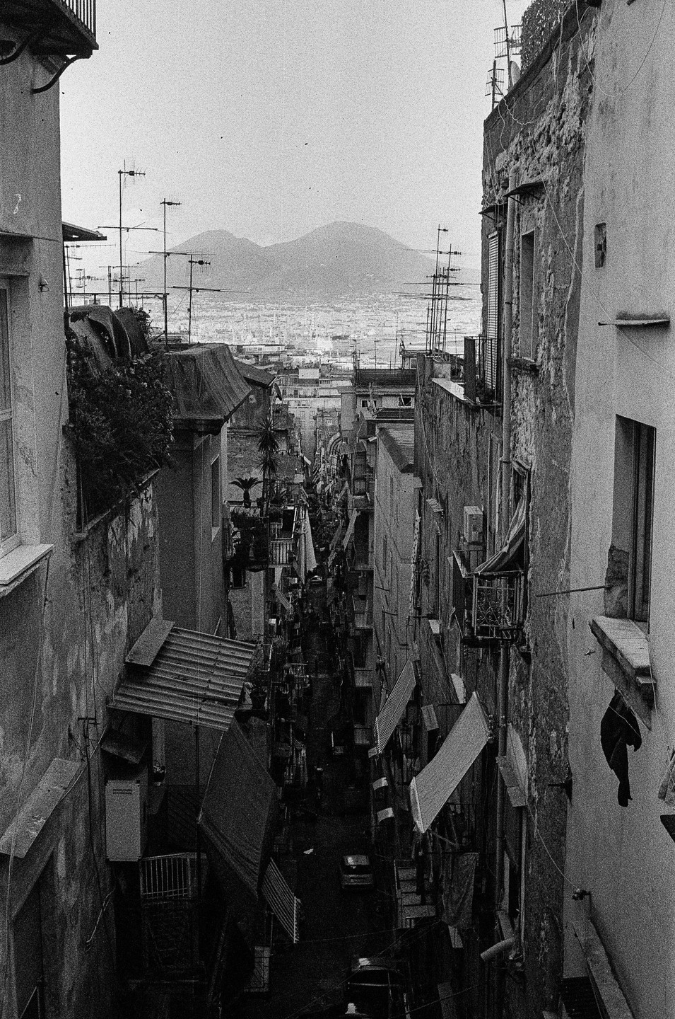 A narrow street in Naples is visible between tall buildings, with mountains and a city in the distance.