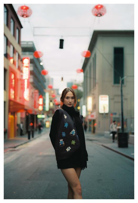 A woman stands in the middle of a street lined with buildings and red lanterns overhead.