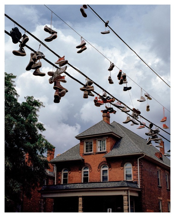 Many pairs of shoes hang from power lines above a brick house with a porch.