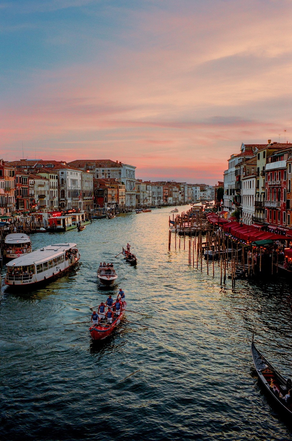 Boats and gondolas travel along a canal lined with buildings under a pink and blue sky at dusk.