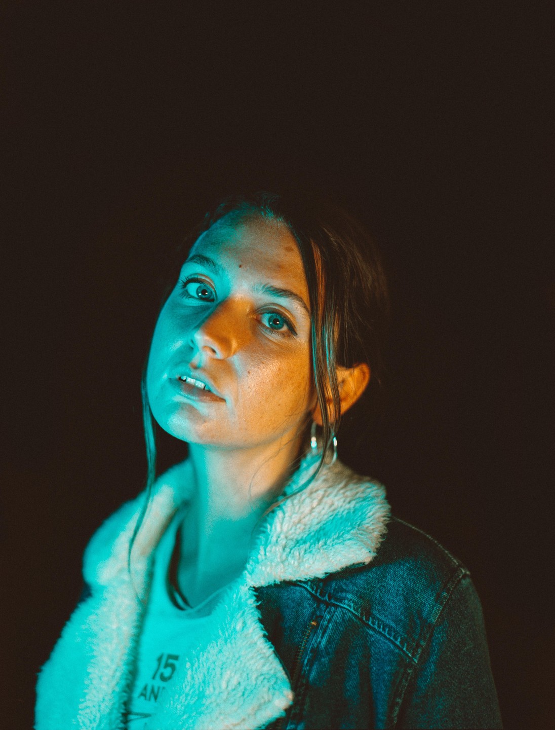 A woman wearing a denim jacket with a fleece collar looks up at the viewer, illuminated by blue and orange light.