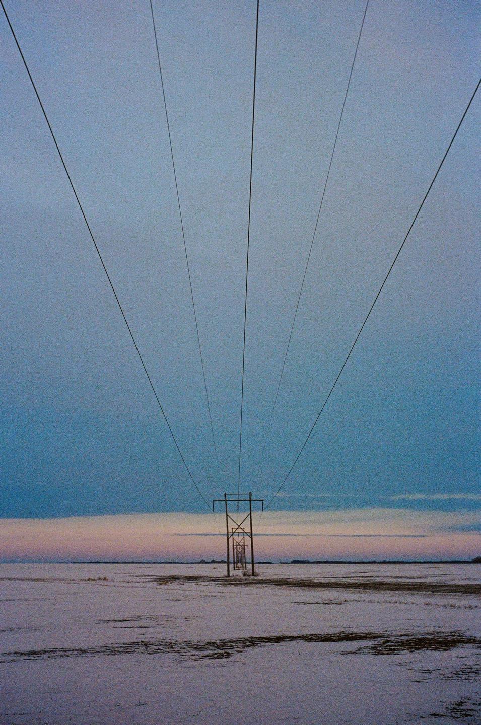 A power line stretches across a snowy field under a blue and pink sky.