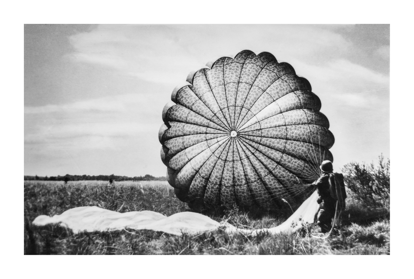 A paratrooper stands next to a partially deflated parachute in a grassy field under a cloudy sky.