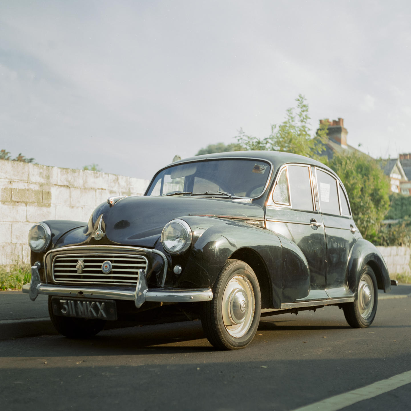 A black Morris Minor car is parked on the side of a street with a stone wall and houses in the background.