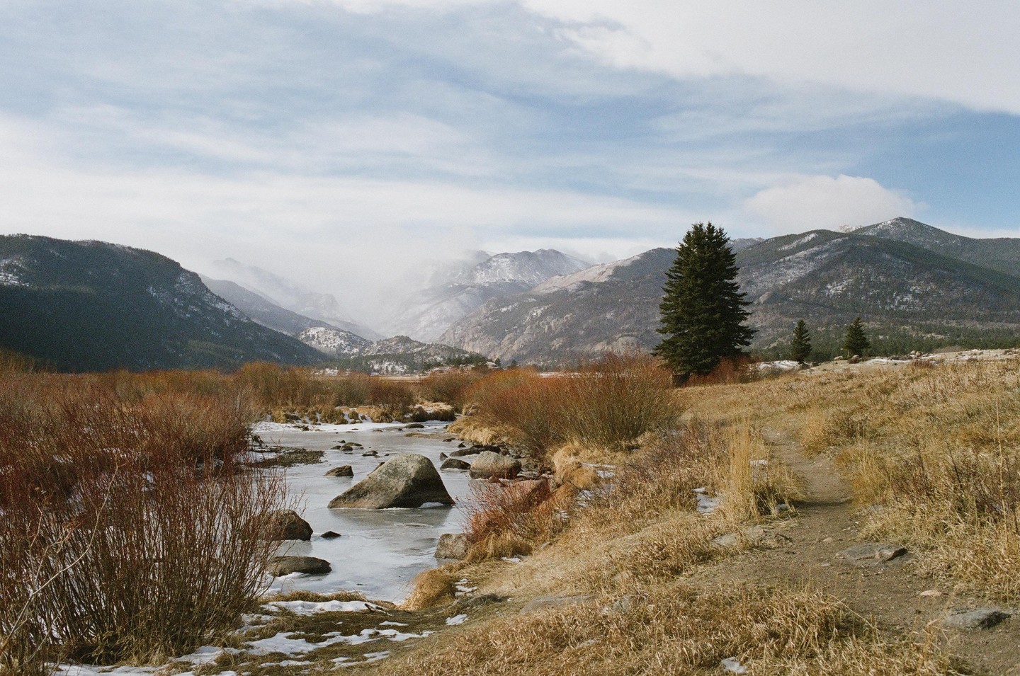 A partially frozen river flows through a valley with snow-capped mountains and pine trees under a cloudy sky.