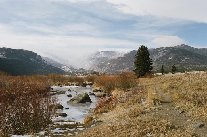 A partially frozen river flows through a valley with snow-capped mountains and pine trees under a cloudy sky.