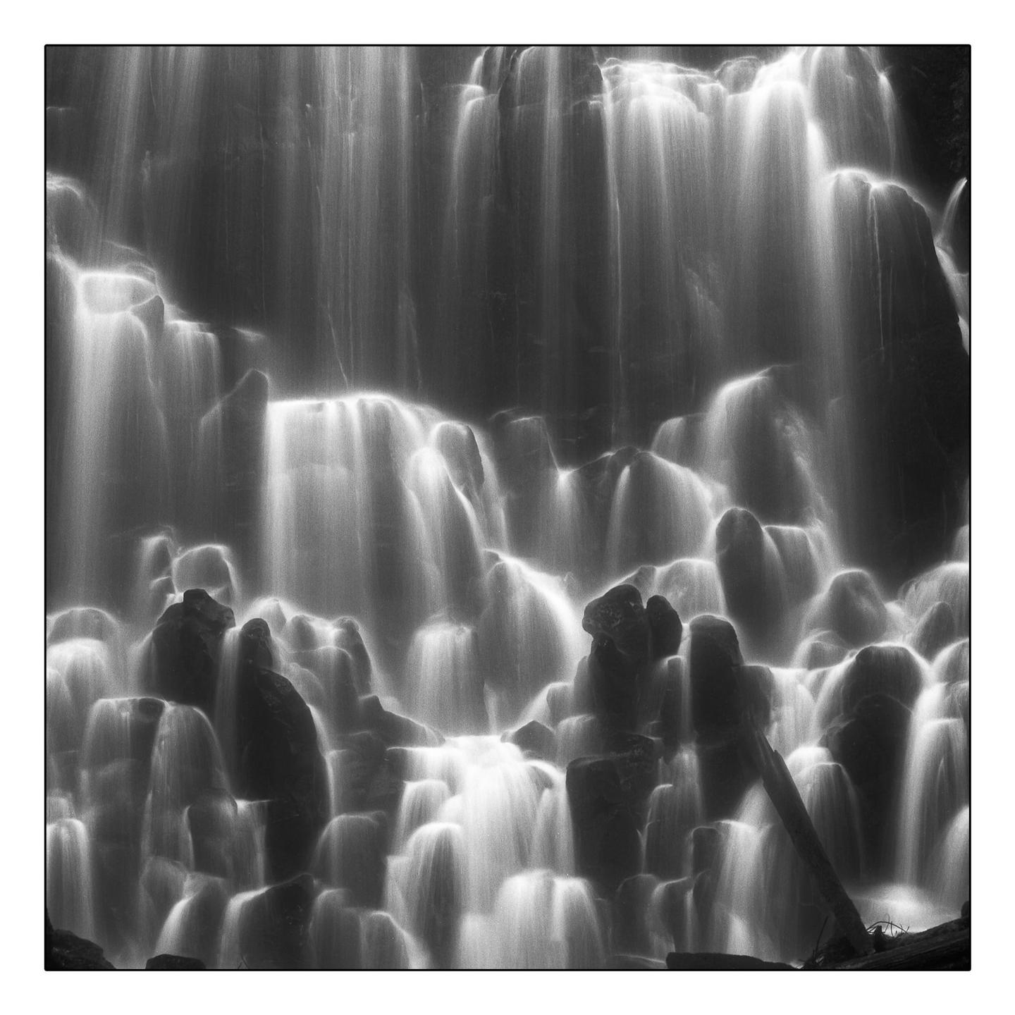 A black and white photograph captures a waterfall cascading over rocks, with the water appearing smooth due to a long exposure.