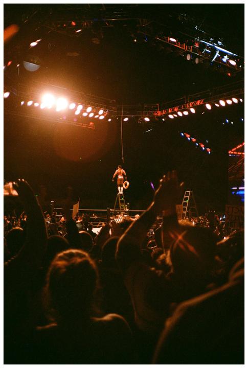 A wrestler stands on a ladder in a brightly lit arena, surrounded by a cheering crowd.