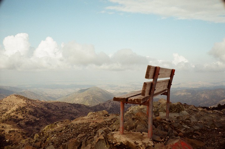 A wooden bench with red legs sits on a rocky mountain overlooking a valley under a cloudy sky.