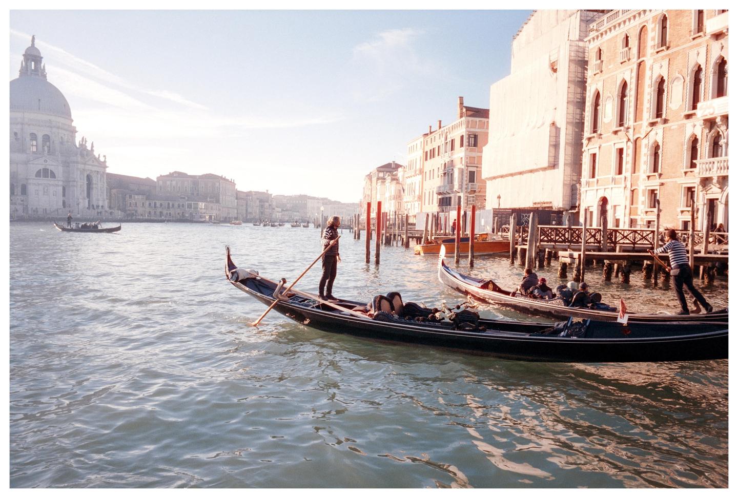 Two gondolas with gondoliers and passengers are on a canal in Venice with buildings and a large domed church in the background.