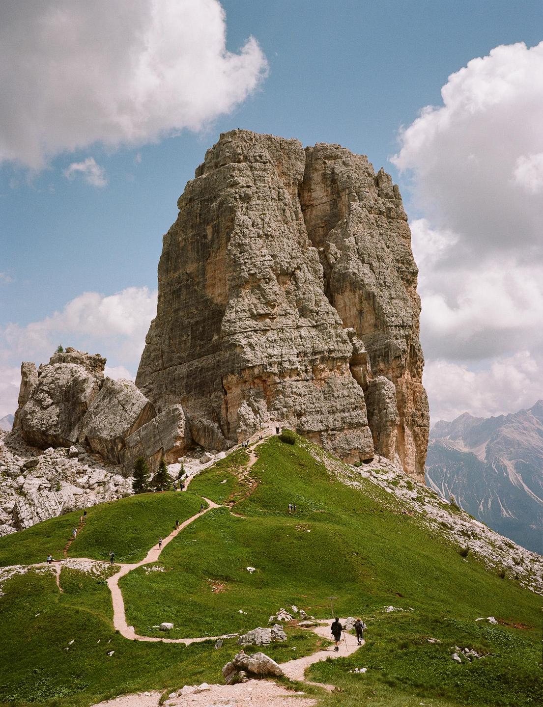 A winding dirt path leads up a grassy hill towards a towering rock formation under a cloudy sky.