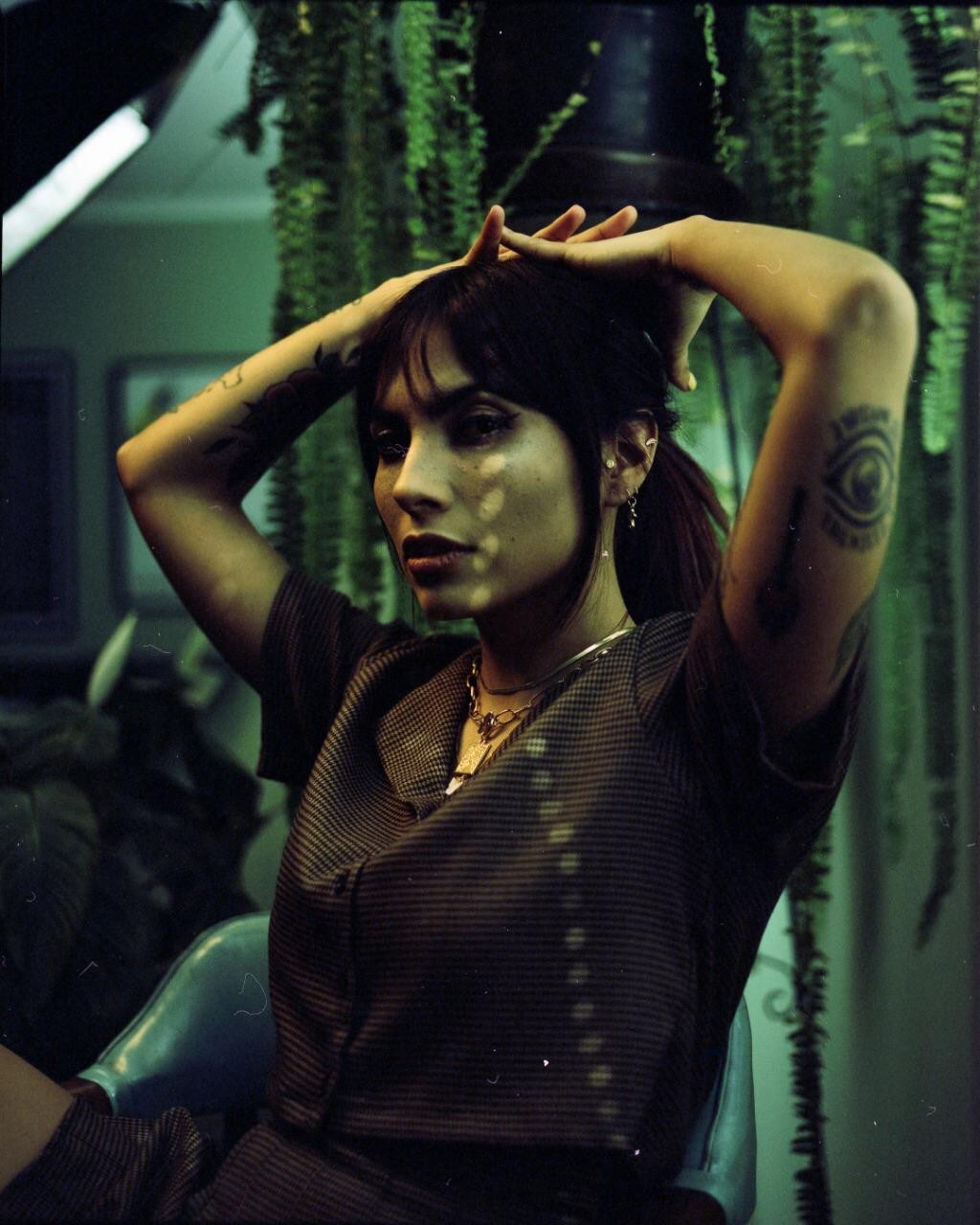 A woman with dark hair and tattoos poses with her hands above her head, illuminated by dappled light, in a room with hanging plants.