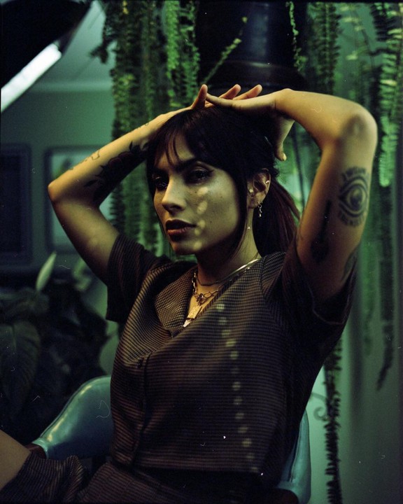 A woman with dark hair and tattoos poses with her hands above her head, illuminated by dappled light, in a room with hanging plants.