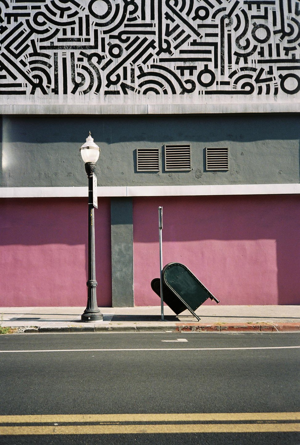 A tilted green mailbox stands on a sidewalk next to a street light, with a pink wall and a patterned mural above it.