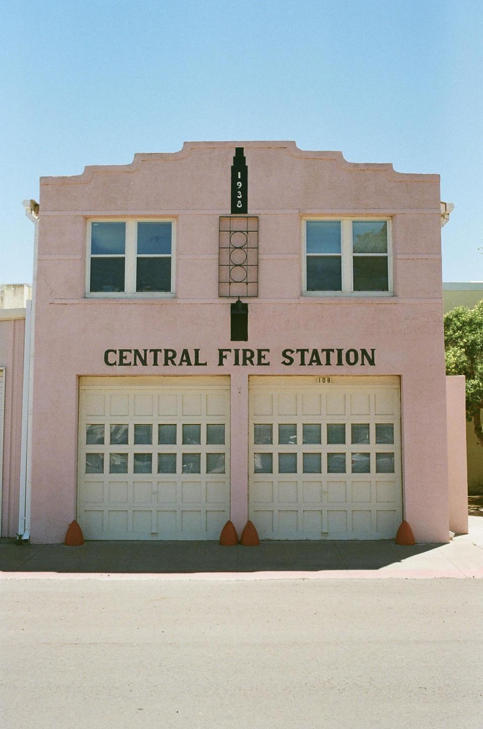 A pink building with "CENTRAL FIRE STATION" written on it has two garage doors and two windows.