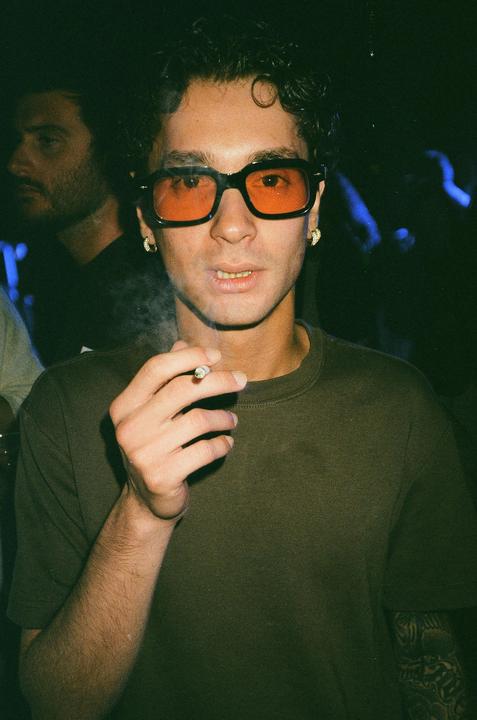 A man wearing orange-tinted glasses and a dark green shirt smokes a cigarette in a dimly lit room.