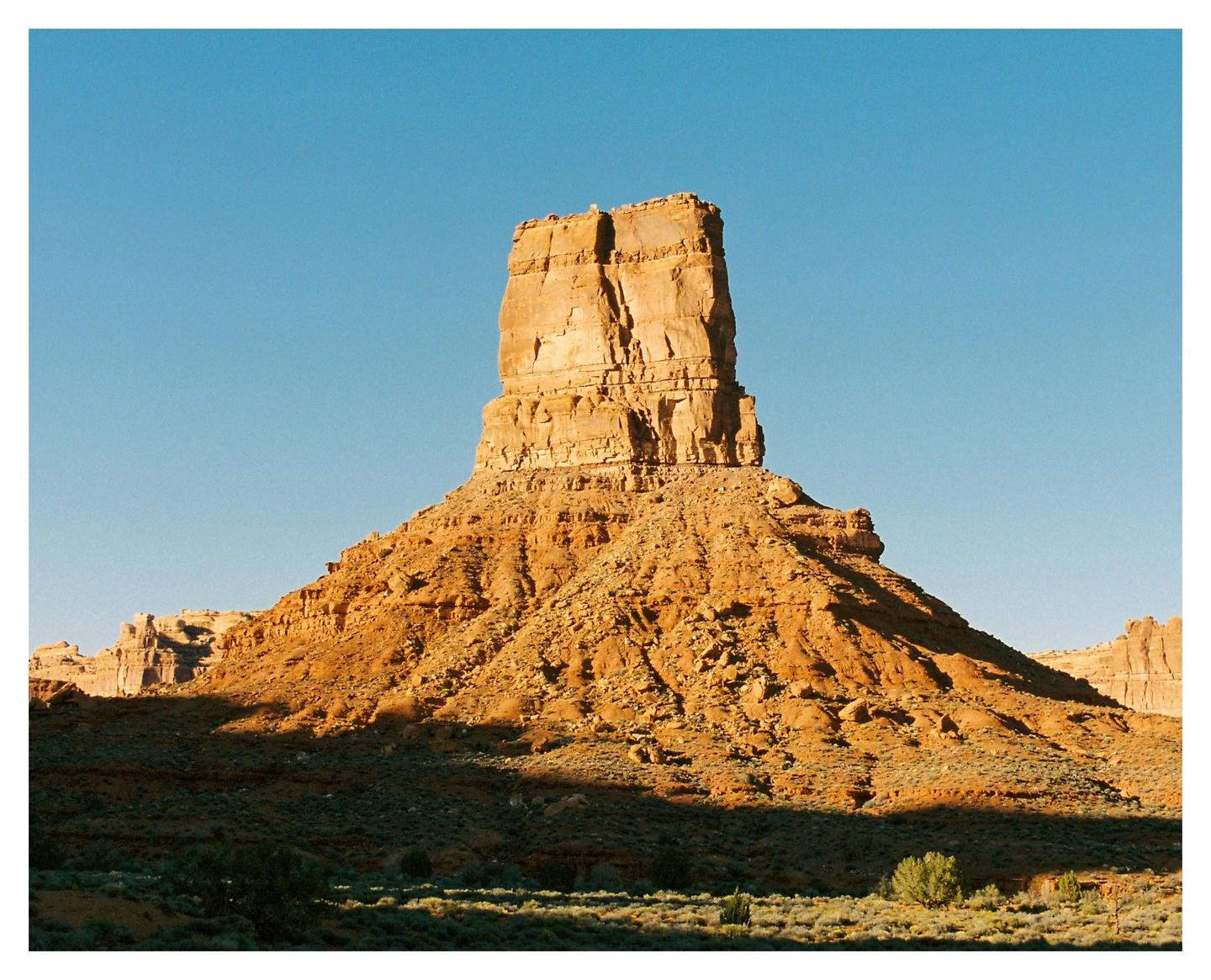 A large rock formation stands tall against a clear blue sky in a desert landscape.