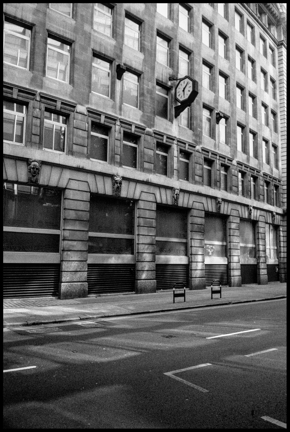 A large building with a clock on its facade stands next to a street with two benches.