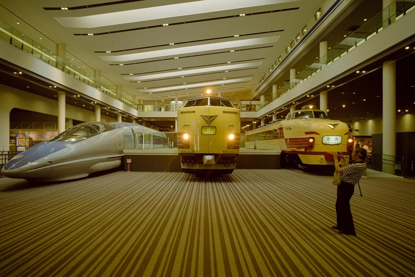 Three trains are on display in a large hall with a woman taking a photo of one of them.