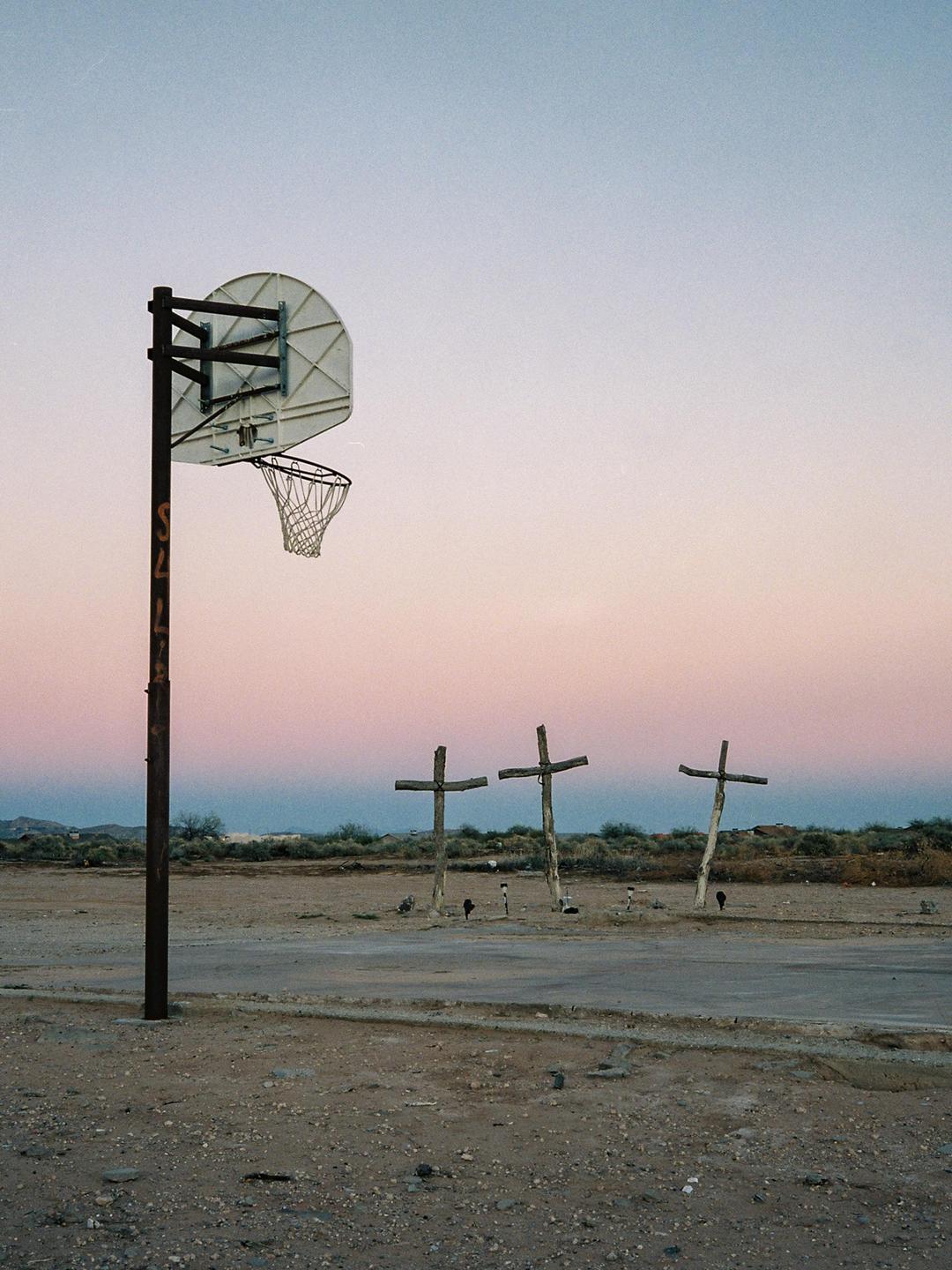 A basketball hoop stands in a desert landscape with three wooden crosses in the distance under a colorful sky.