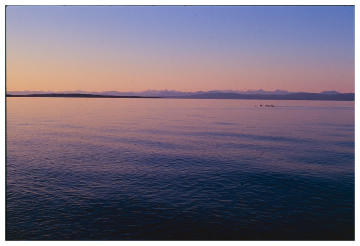 A body of water with a distant landmass and mountains under a colorful sky at dusk.