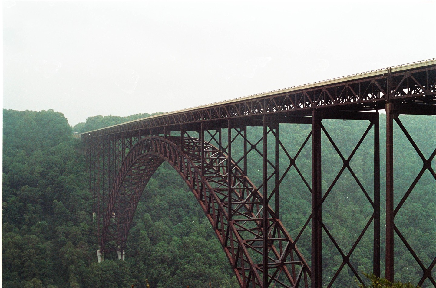 A long, arched bridge spans a wide gorge filled with green trees under a bright, overcast sky.