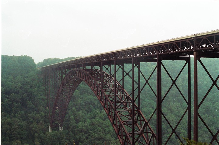 A long, arched bridge spans a wide gorge filled with green trees under a bright, overcast sky.
